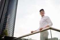 Smiling young businessman in elegant outfit smiling and looking away while standing near railing on street of modern city — Stock Photo