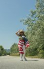 Glückliche Frau, die mit einer amerikanischen Flagge auf einer einsamen Straße spaziert und feiert. Besonderer Tag zur Feier des 4. Juli — Stockfoto