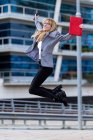 Блондинка в очках и сером пиджаке с красной сумкой и прыжками с телефона перед современным зданием — стоковое фото