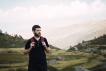 Adult bearded man with backpack standing in green rocky valley and looking away, Spain — Stock Photo