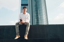 Lächelnder junger Mann mit Smartphone sitzt an Wand gegen modernen Wolkenkratzer — Stockfoto