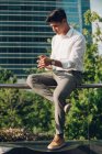 Bello giovane ragazzo in abito elegante seduto su ringhiera sulla strada della città e utilizzando smartphone — Foto stock