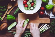 De dessus assortiment de légumes mûrs rouges et verts et les mains de la personne hacher des concombres sur planche à découper en bois
. — Photo de stock