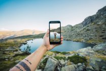 Mano de hombre tatuado sosteniendo teléfono inteligente como tomando la foto de la mujer en las rocas de pie en la orilla del lago de montaña - foto de stock