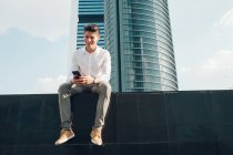 Усміхнений молодий чоловік зі смартфоном сидить на стіні проти сучасного хмарочоса — стокове фото