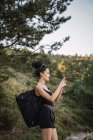 Vista lateral de morena con mochila de pie en la naturaleza salvaje y tomando fotos con smartphone, España - foto de stock