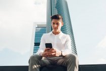 Молодий чоловік зі смартфоном сидить на стіні проти сучасного хмарочоса — стокове фото