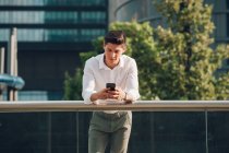 Jovem empresário com smartphone apoiando-se em trilhos em frente ao edifício moderno — Fotografia de Stock