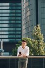 Jovem empresário sorridente com smartphone apoiado em trilhos na frente do edifício moderno — Fotografia de Stock