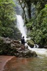 Rückansicht eines Mannes mit Rucksack, der auf einem nassen Felsbrocken steht und einen fantastischen Wasserfall im Regenwald in Costa Rica betrachtet — Stockfoto