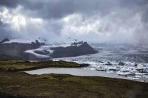 Green shore and snowy cliffs near ice lake in cloudy weather in Iceland — Stock Photo