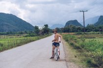 Uomo senza maglietta seduto in bicicletta e guardando indietro sulla strada rurale in collina. — Foto stock