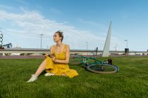 Femme élégante en robe jaune fluide et baskets assis sur la pelouse avec vélo et petit ordinateur portable — Photo de stock