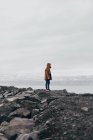 Persona anónima en abrigo de pie en la costa de rocas grises con agua brumosa en el fondo, Islandia. - foto de stock