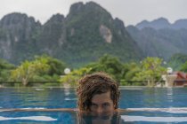 Привлекательный молодой человек смотрит в камеру и плавает в бассейне на фоне зеленых холмов. — стоковое фото