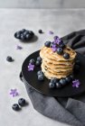 Stack of appetizing tasty crumpets with blueberries and purple flowers on black plate on grey background — Stock Photo
