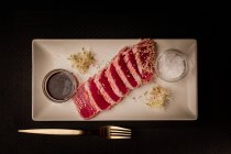 Сырой нарезанный стейк из тунца на блюдечке с соусом на черном фоне — стоковое фото