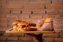 Mano humana sosteniendo sabrosas hamburguesas y papas fritas en bandeja de madera - foto de stock