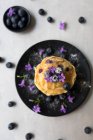 Stack of appetizing tasty crumpets with blueberries and purple flowers on black plate on grey background — Stock Photo