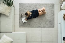 Junge Frau kuschelt auf Teppich im Wohnzimmer, liest Buch und trinkt Kaffee — Stockfoto