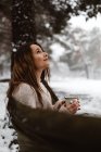 Vista lateral de la mujer joven bastante soñadora sentada en la hamaca y teniendo bebida caliente en la naturaleza del invierno. - foto de stock