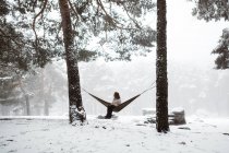 Vista lateral da jovem mulher muito sonhadora sentada na rede e tendo bebida quente na natureza de inverno. — Fotografia de Stock