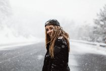 Vista lateral da mulher bonita de pé e olhando para a câmera na estrada em queda de neve. — Fotografia de Stock