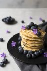 Montón de sabrosos bocadillos apetitosos con arándanos y flores de color púrpura en el plato negro sobre fondo gris - foto de stock
