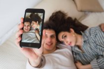 Joyeux jeune couple prenant selfie avec smartphone sur le lit — Photo de stock