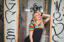 Giovane modella bionda in denim e t-shirt colorata in piedi provocatoriamente in bagno abbandonato con graffiti a parete. — Foto stock
