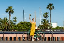 Женщина в летнем жёлтом платье, опирающаяся на велосипед и удерживающая выпивку — стоковое фото