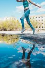 Faceless shot of slim girl in denim jumping high above puddle reflecting blue sky and city. — Stock Photo