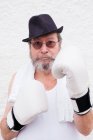 Hombre adulto en sombrero y guantes de boxeo blancos en la pared áspera. - foto de stock