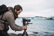 Side view of man in outwear with backpack setting photo camera on tripod to take photo of beautiful cold seascape. — Stock Photo