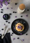 Pilha de apetitosos saborosos crumpets com mirtilos e flores roxas na placa preta no fundo cinza — Fotografia de Stock