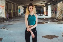 Young beautiful girl in top and jeans standing among broken columns in abandoned building looking at camera. — Stock Photo