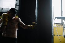 Боксер без рубашки в перчатках стоит и бьет грушу во время тренировки. — стоковое фото