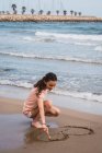 Teen girl squatting and painting with stick on sand on seashore — Stock Photo