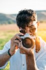 Красивий молодий хлопець в сонцезахисних окулярах весело посміхається і тримає ретро фотоапарат, стоячи на розмитому тлі дивовижної сільської місцевості — стокове фото