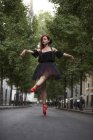 Bailarina cabeça vermelha com tutu preto e dicas de balé vermelho dançando na rua com árvores no fundo . — Fotografia de Stock