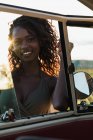 Mulher afro-americana encantadora sorrindo e olhando para a câmera através da janela do carro vintage enquanto passa o tempo na natureza no dia ensolarado — Fotografia de Stock