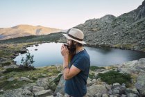 Junger Mann mit Hut steht in der Nähe eines Sees in den Bergen mit Becher — Stockfoto
