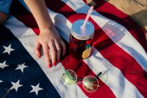 Schnappschuss einer Frau, die mit Getränken und Sonnenbrille auf amerikanischer Flagge im Sonnenlicht sitzt. — Stockfoto