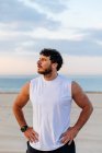 Homme positif en vêtements de sport avec les mains à la taille tout en se tenant sur la plage de sable fin pendant le coucher du soleil — Photo de stock