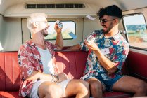 Deux jeunes gens joyeux riant et jetant des cartes à jouer assis sur le siège passager du van rétro pendant le voyage dans la nature — Photo de stock