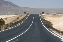 Clean straight road with signs on dry sandy plain with hills and mountains, Canary Islands — Stock Photo