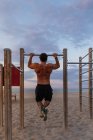 Вид ззаду м'язистого хлопця, що виконує підтяжки на барі під час заходу сонця на піщаному пляжі — стокове фото