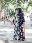 Attractive brunette woman in dark dress standing on street and smelling fresh beautiful flowers on sunny street — Stock Photo