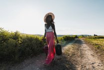 Femme afro-américaine à la mode en chapeau portant une valise et marchant sur la route rurale en été — Photo de stock