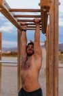 Бородатый мужчина без рубашки поднимается по деревянной лестнице во время тренировки на пляже — стоковое фото
